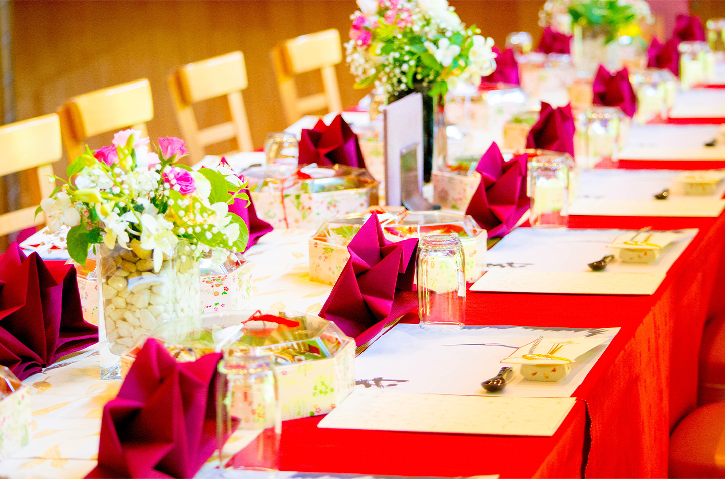 赤いテーブルクロスにワイン色のナプキンを配した、祝い席向けの華やかな宴会テーブルセッティング(花の装飾と個別の席準備が整った会場風景)