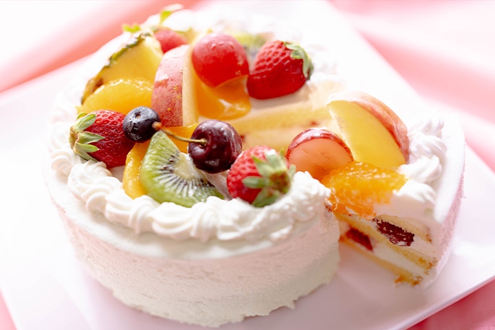 いちごやキウイ、桃などのフルーツをたっぷり飾ったデコレーションケーキを一切れ取り分けたお祝い用ホールケーキの写真