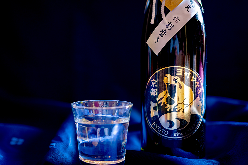 与謝娘酒造の日本酒ボトルとグラス。京都与謝野町産コシヒカリ使用、六割磨き表示。青い背景に金色のカエルのラベルが映える。