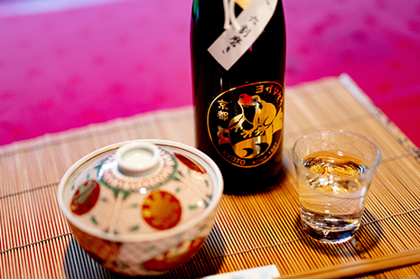 京都 与謝野町 与謝娘酒造の純米吟醸 コシヒカリ使用日本酒と和食の膳。金のカエルラベルが特徴。竹のランチョンマットに酒瓶とおちょこ、蓋付き椀を並べた料理写真。